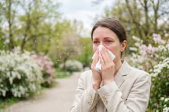 Femme souffrant d'allergies saisonnières dans un parc au printemps, se mouchant entourée de pollen et d'arbres en fleurs
